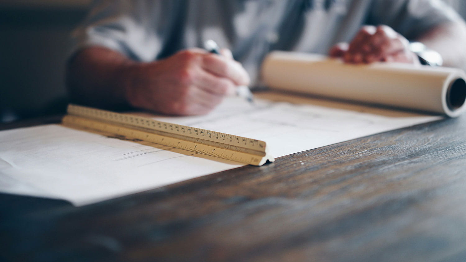 Person drafting plans at a wooden table, writing on large sheets of paper with a folded ruler and rolled blueprint in the foreground.